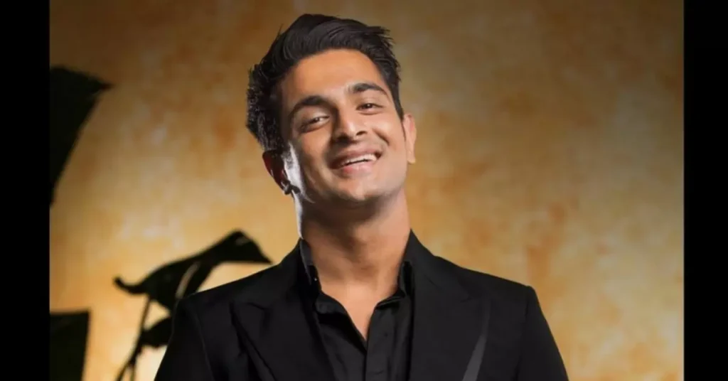 The image shows a smiling man in a black suit and tie with a black shirt underneath. The man is standing in front of a dark background with a black wall behind him. The man is wearing a smile on his face, which suggests that he is happy or feeling positive about something.